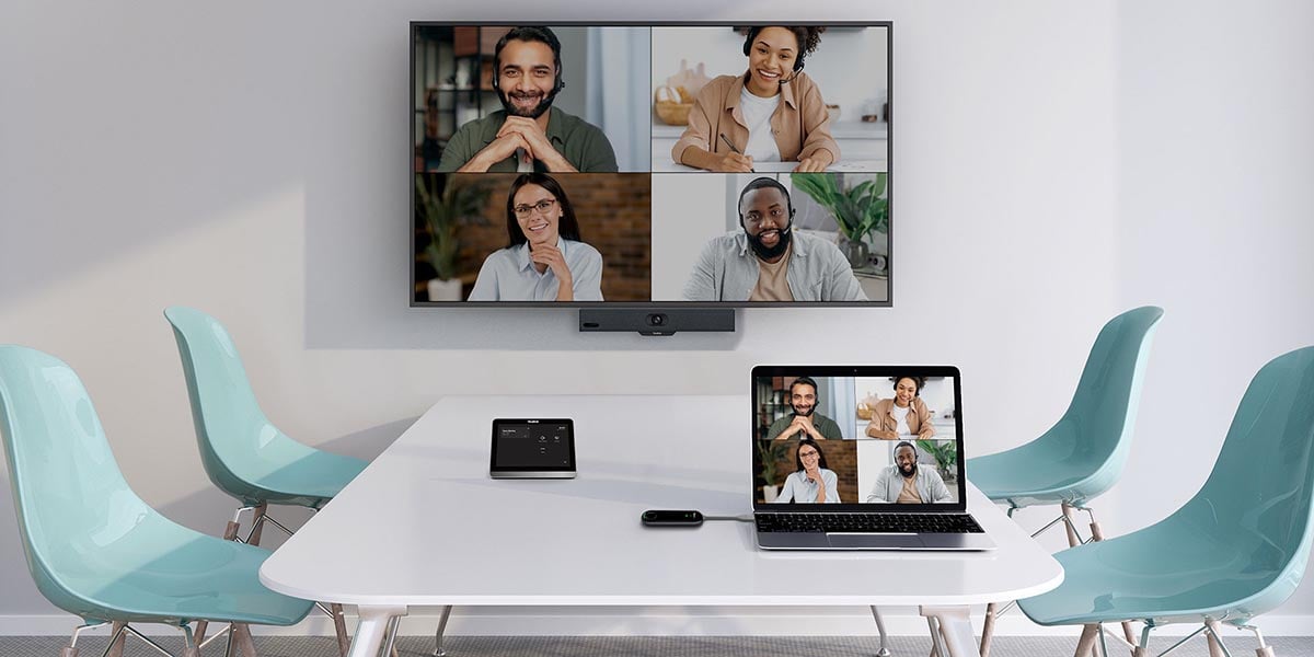 ein Bildschirm mit 4 Personen die an einer Videokonferenz teilnehmen. Vor dem Bildschirm steht ein Tisch mit Laptop und vier Stühlen